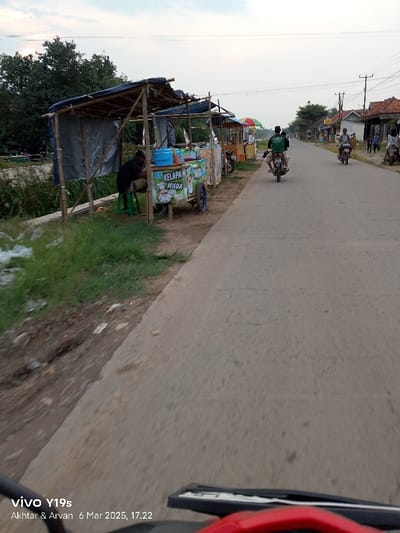 The image shows a street scene in Kecamatan Gabuswetan, Indonesia, located at latitude -6.438 and longitude 108.024. It is a paved road with a line of food stalls on the right side. The stalls are made of bamboo and have thatched roofs. A man is sitting on a stool in front of the closest stall, which is selling "Kelapa Muda," which translates to "Young Coconut." There are other people in the background, including a man on a motorcycle and a few people walking. The environment is rural with trees and houses on either side of the road. The sky is overcast. The picture was taken from a moving vehicle facing the road. The bottom of the image includes a red handlebar of a motorcycle and a "vivo Y19s" logo. The date and time are "6 Mar 2025, 17:22." The photo is slightly blurred, likely due to the movement of the camera.