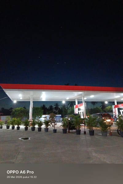 The image shows a gas station in Sawang, Indonesia, at night. The red and white canopy of the station is illuminated by bright lights. Several cars are parked under the canopy, and people are visible. Potted plants line the front of the station. The sky is dark, with a few stars visible. The pavement is wet, reflecting the lights from the station. A timestamp indicates the photo was taken on May 26, 2026, at 19:22.