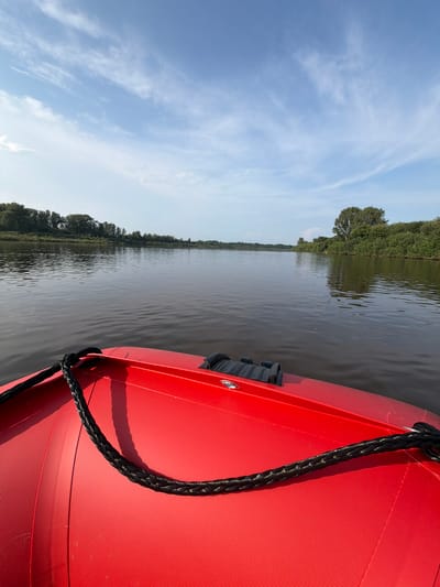 A first-person perspective shot shows the front of a red inflatable boat on a wide, dark river under a bright blue sky with wispy white clouds. The river extends into the distance, bordered by a lush green treeline on both banks. The rope on the front of the boat is black and braided. The location is Kazaevo, Russia.