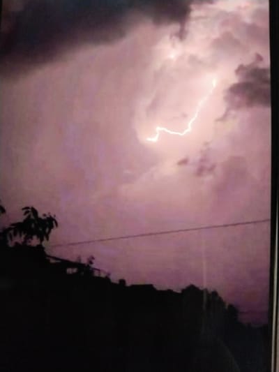 The image captures a dramatic sky illuminated by a lightning strike. Dark, moody clouds dominate the upper portion of the frame, with a bright, jagged line of lightning splitting through them. The overall color palette is a deep purple and pink, suggesting twilight or a storm-laden evening. In the lower portion of the image, a silhouetted landscape is visible. This includes the dark outlines of trees and what appear to be rooftops, indicating a residential or semi-urban area. A thin, horizontal line, likely a utility wire, stretches across the middle ground. The scene is devoid of people and any specific identifiers of Tinchlik, Uzbekistan.