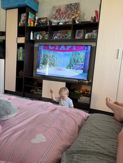 The image depicts a living room scene inside an apartment in the city of Tsentralny, Russia. A young child with blonde hair is sitting on a bed covered with pink and white striped bedding, looking towards a television screen that is displaying a scene from an animated movie. The child is wearing a white t-shirt and has a pacifier in their mouth. On the bed, there are stuffed animals.
The television is mounted on a shelf unit that also holds various decorative items, books, and framed photographs. To the right of the television, a pair of adult feet with bright yellow toenail polish are visible. The room appears to be a typical residential interior, with wallpaper on the walls and furniture that includes a large wardrobe. The overall impression is one of a casual, domestic setting.