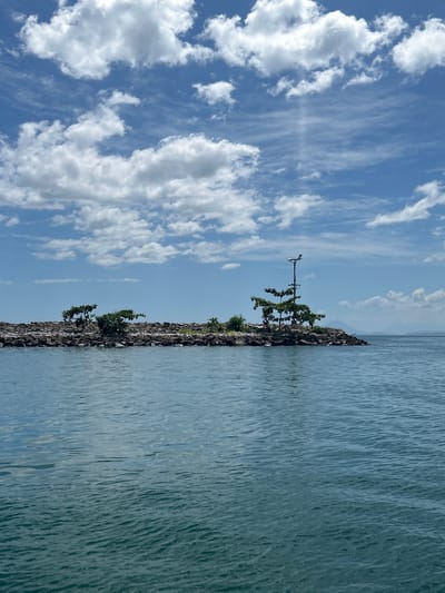 A serene seascape of the waters surrounding the city of Tidore Islands, Indonesia, is depicted under a bright, partly cloudy sky. The foreground is dominated by calm, blue-green water with gentle ripples. In the mid-ground, a rocky islet juts out from the water, densely covered with dark stones and scattered with small green trees and shrubs. A single, tall lamppost stands on the islet, adorned with a light fixture at its apex. The background reveals a distant, hazy landmass on the horizon and a vast sky filled with fluffy white clouds against a clear blue backdrop. The scene conveys a sense of tranquility and natural beauty.