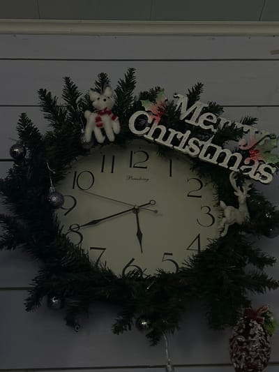 The image shows a close-up, head-on, vertical shot of a Christmas-decorated clock against a light blue wooden plank wall. The clock face is white with black Arabic numerals. The hour hand is pointing between 8 and 9, and the minute hand is pointing to the 10. A small inscription "Baisheng" is visible below the 12.

The clock is surrounded by a circular wreath made of dark green artificial pine branches. Several silver baubles are attached to the wreath. A white plush reindeer with a red and white striped scarf stands near the top center of the wreath. Silver glittery text spelling "Merry Christmas" is affixed to the wreath. A small white reindeer figure is positioned on the right side of the wreath, facing left. A decorative pine cone with white frosting and a red bow is hanging from the bottom right of the frame.

The lighting in the image is dim, casting shadows and highlighting the reflective surfaces of the baubles and the "Merry Christmas" text. The wall behind the clock has a horizontal plank texture. There are no people visible in the image. The scene does not provide any information that can be used to determine its location within the city of Appany, Russia.