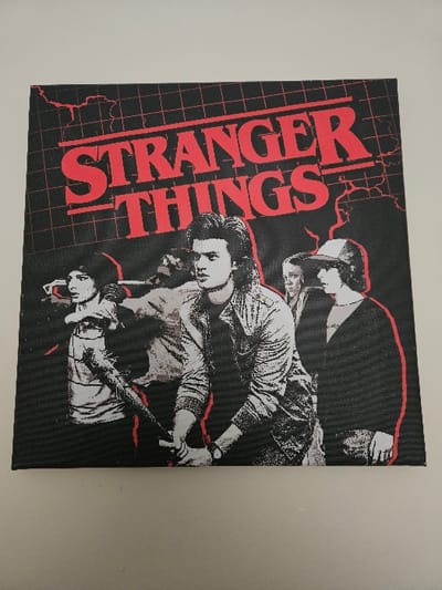 A square canvas print featuring promotional artwork for the television series "Stranger Things" is displayed flat against a plain, light-colored wall. The print itself has a black background with a red grid pattern and red lightning bolt outlines. The title "STRANGER THINGS" is prominently displayed in large red capital letters across the top. Below the title, five figures are depicted in a monochromatic style with red accents. The central figure, a male with long, feathered hair, wears a denim jacket and holds a spiked baseball bat with both hands, positioned defensively. To his left, a male wearing glasses extends his left arm forward, holding an unseen object. A partially obscured figure wearing a cap is positioned behind the central male to the left. To the central male's right, a female figure with dark hair looks forward intently, and further to the right, another male figure wearing a cap is visible. The figures are grouped together in a confrontational or defensive stance. This item is located within the Cherwell District, United Kingdom.