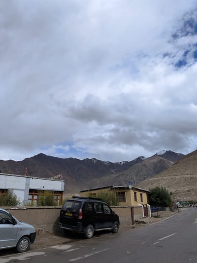 The image depicts a scene in the city of Khardung, India, on a cloudy day. In the foreground, a black SUV is parked on the side of a paved road. To the left of the SUV, a silver car is also parked. Behind the cars, there is a low wall made of concrete and stone. Beyond the wall, a white building with dark trim and windows is visible, adorned with colorful prayer flags. Further back, a yellow building with similar windows is present. The background is dominated by a mountain range, with some peaks appearing snow-capped. The sky is filled with a mixture of white and gray clouds.