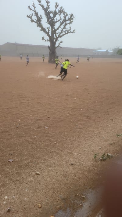 A group of people, presumed to be playing soccer, are in an open, dirt field near a large tree with bare branches. The scene is hazy, possibly due to fog. The location is the city of Jogana, Nigeria. The players are wearing either yellow or white shirts. One player is kicking a soccer ball, creating a small cloud of dust. A building can be seen in the background.