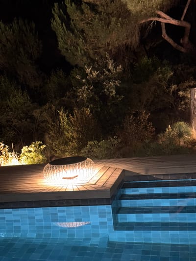 A low-angle, night time photograph captures a serene outdoor setting adjacent to a swimming pool. In the foreground, the turquoise water of the pool is visible, with its tiled surface reflecting the ambient light. To the right, a set of tiled steps descends into the pool.
A wooden deck occupies the mid-ground, featuring a woven, orb-shaped lamp that emits a warm, inviting glow. The lamp is positioned on the edge of the deck, near the steps. Beyond the deck, dense foliage and trees form a dark, natural backdrop. Some of the plants are illuminated by spotlights, casting a warm light on their leaves and branches. The overall atmosphere is tranquil and inviting, suggestive of a peaceful evening in an outdoor living space.
The image is identified as being located in Grândola, Portugal.