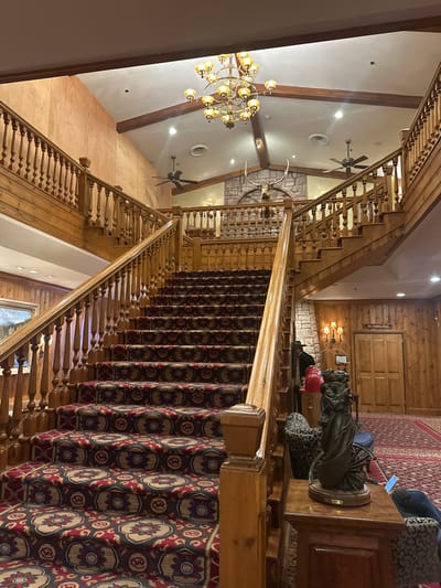 An interior view looking down a grand wooden staircase covered with patterned carpet. A large chandelier hangs from the ceiling, which also features ceiling fans and wooden beams. A stag's head adorns a wall. The staircase ascends to a landing with wooden walls and a doorway. A statue of a kneeling person is near the top of the stairs.