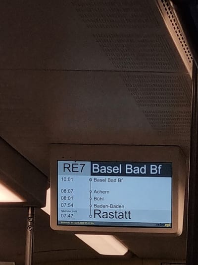 A digital display shows a train schedule with the heading "RE7 Basel Bad Bf." The display indicates the train's route, showing stops at Basel Bad Bf, Achern, Bühl, Baden-Baden, and Rastatt. Scheduled departure times are listed next to each stop. The display notes the next stop is at Rastatt, scheduled for 07:47. The date shown at the bottom of the display reads "Wednesday, 01 April 2028 17:41 Uhr". The location is not stated.