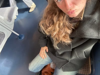 The scene is located within a public transport vehicle in Bondy, France. The image is taken from a high angle, focusing on a young adult female seated in the foreground. She has long, wavy, light brown hair and blue eyes, captured with pouting lips in a selfie-style pose. She wears a dark overcoat, a collared patterned sweater underneath, and light blue denim jeans. Two rings are visible on her left hand. Her right foot, partially visible, shows a light-colored patterned shoe.

To her left, the back of a light-colored public transport seat with a patterned fabric is visible, along with a metal support pole anchored to the dark blue floor. Another individual's lower legs and feet are partially visible in the upper left background; this person wears light grey trousers and cream-colored, pointed-toe ankle boots. The floor is uniformly dark blue, and a circular blue pole base is centered in the mid-left of the frame.