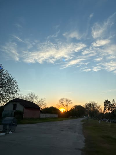 The image, captured in Glojene, Bulgaria, depicts a serene sunset over a landscape featuring a cemetery. The sky dominates the upper portion of the frame, transitioning from a clear blue on the left to warm oranges and yellows towards the setting sun on the right horizon. Delicate, wispy white cirrus clouds are scattered across the blue sky, with subtle blue lens flares visible.
The sun is prominently featured on the right, glowing brightly just above the horizon, partially obscured by the silhouettes of trees. Some trees appear to have bare branches, while others show early foliage. Below the horizon, in the lower right, a small building with a dark, possibly red-brown tiled roof and lighter walls is nestled among more trees.
A paved or dirt road curves from the lower right towards the mid-ground, flanked by areas of green grass. In the mid-right distance, several light-colored, upright structures resembling gravestones or monuments are visible among the trees and grass, strongly indicating the scene is set within or borders a cemetery. The time of day is clearly sunset or late afternoon. The weather appears clear with high-altitude clouds. No people, discernible activity, or visible text are present in the peaceful, contemplative scene.
