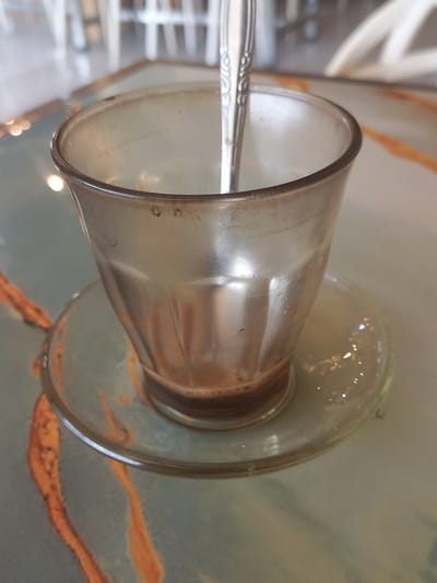 The image shows a glass cup with a brown liquid inside, resting on a saucer on a table with a patterned surface. A spoon is inserted into the cup. The location in the city of RawangItiek, Indonesia is not confirmed in the image.