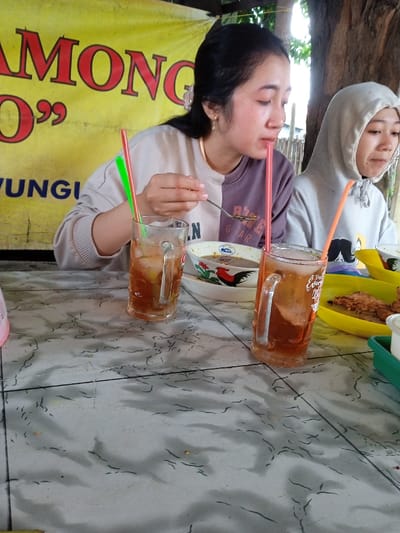 Two women sit at a table in what appears to be an outdoor eatery. The woman in the foreground has a two-toned sweatshirt, eats from a bowl, and drinks from a glass with a straw. Beside her, the second woman wears a hooded sweatshirt. The table is tiled and holds the women's food, drinks, and condiments. A sign is partially visible behind the women. A tree trunk is in the background on the right.