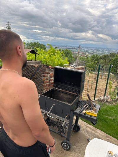 Here is a description of the image:

A somewhat rotated, wide-angle shot shows a man grilling outdoors. The man is fair-skinned with short hair and a gold chain, and is wearing black shorts. He stands next to a black barbecue grill. A brick structure supports something that has "SKYFORM" printed on it. Grilling tools are on the grill. A white plastic table is to the side. A view of a city, possibly Brestnik, Bulgaria, extends into the distance under a cloudy sky. A fence is visible in the mid-ground. The setting appears to be a patio or yard.