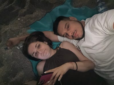 A couple is lying on a turquoise blanket on the ground, which appears to be sandy or rocky, in El Parador de las Hortichuelas, Spain. The man, on the right, is wearing a white t-shirt and has dark hair and a mustache. He is looking towards the camera. The woman, on the left, has long dark hair and is wearing a dark-colored top. She is also looking towards the camera, holding a pink smartphone in her hands. Both individuals appear relaxed and are lying down. The background is dark and out of focus, suggesting it is nighttime or a dimly lit environment.