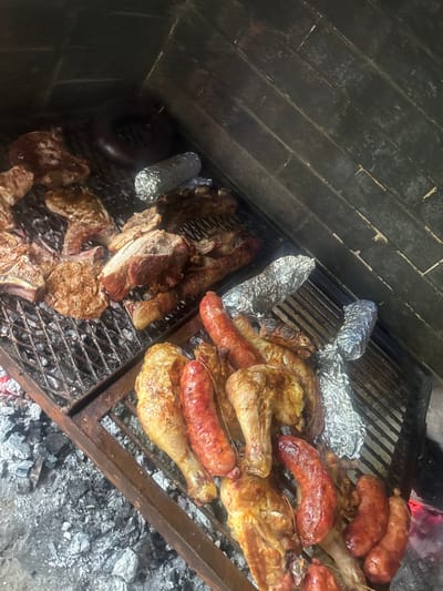 A high-angle shot captures a vibrant Argentine asado (barbecue) in progress, with an array of meats cooking over hot coals within a traditional brick parrillero structure. The scene is illuminated by the glow of the embers and ambient low light, suggesting it is evening or night.

The main subjects are the various cuts of meat arranged across two large metal grills. On the left side, substantial pieces of beef, including what appear to be ribs and steaks, are seen browning on the grates. Interspersed among the meats are several items wrapped in aluminum foil, likely vegetables or other slow-cooked accompaniments.

The right grill section holds a collection of chicken pieces, specifically drumsticks and thighs, alongside multiple large, reddish-brown sausages (chorizos), all appearing to be well-cooked with a tempting golden-brown hue. More foil-wrapped items are also present on this side.

Below the grills, a bed of white ash and glowing red embers indicates the source of heat. The sturdy, dark brickwork of the parrillero forms the background on the left and top of the image, while the ash-covered ground is visible on the lower right.

The image depicts a quintessential grilling event, characteristic of the cultural practices in Longchamps, Argentina, where an asado is a significant social gathering. No people are visible in the frame, and there is no discernible text. The focus is entirely on the plentiful food and the cooking process, evoking a sense of warmth, good food, and convivial atmosphere.