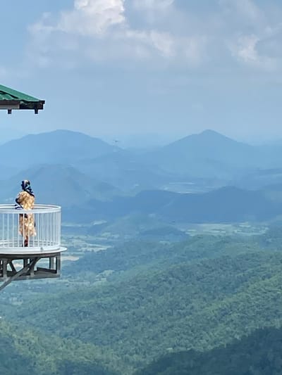A person stands on a white, circular observation deck, looking out at a vast expanse of mountains. The person is wearing a light-colored dress with a patterned design and a headscarf. The observation deck is supported by a wooden structure and has a railing. A portion of a green-roofed structure is visible in the upper left corner. The background is a series of layered, hazy blue mountains, with the closest ones covered in dense green forest. The sky is a pale blue with scattered white clouds. The scene appears to be at a high elevation, offering a panoramic view of the natural landscape. The location is identified as the observatory in Nam Hải, Vietnam.