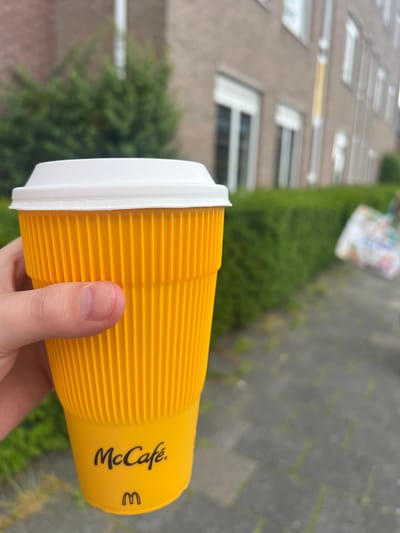 A hand holds a bright yellow McCafe branded paper cup with a white lid. The cup has a ribbed texture. The McCafe logo and a smaller "M" logo are visible in black text on the cup. The background is out of focus, showing a brick building with windows and some greenery, suggesting an outdoor urban setting. The lighting suggests it is daytime. The image is taken from a slightly upward angle.