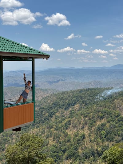 The image captures a vibrant mountainous landscape with a person posing on a viewing platform. The scene is dominated by layers of green mountains stretching into the distance under a bright blue sky dotted with scattered white clouds. The mountains are covered in dense forest, with some patches appearing drier or less vegetated. A plume of white smoke, possibly from a small fire, rises from the trees on one of the closer hills.
In the foreground, a person, appearing to be a young woman, is standing on a balcony or observation deck. She is wearing a striped long-sleeved crop top and denim shorts. She is leaning backward, extending her arms out to her sides, striking a playful or energetic pose as she takes in the expansive view. The structure she is standing on features a green corrugated roof visible on the left, and a wooden-paneled section on the right, framing the mountainous panorama. A dark metal railing is present at the edge of the platform.
The weather is clear and sunny, indicative of daytime. The lush greenery and layered mountains suggest a scenic, possibly remote, natural location in Phan Son, Vietnam. There is no visible text in the image.