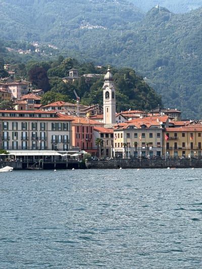 The image depicts a picturesque waterside town nestled against a verdant, tree-covered mountainside. A calm, rippling body of water, likely a lake, dominates the right side of the frame. Along the shore, a series of buildings with terracotta roofs and various colored facades are arranged in a linear fashion. The architecture suggests an European setting, with balconies, shutters, and a prominent church tower with a clock face visible. The buildings appear to be a mix of residential and commercial properties, with some featuring outdoor seating areas.
The mountainside is densely covered with lush green trees, creating a dramatic backdrop to the town. A faint haze is present in the distance, softening the view of the higher slopes. Several small boats are docked along the waterfront, and white buoys are visible in the water. The lighting suggests it is daytime, likely mid-morning or afternoon, with soft shadows. The overall impression is one of a serene and charming location, typical of a lakeside Italian town. No people are clearly discernible in the image.