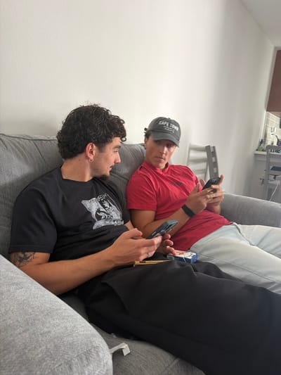 Two young men are shown lounging on a grey upholstered couch in a bright, minimalist indoor setting, likely a living room, in Redondo Beach, United States of America. The room features stark white walls and clear, even lighting, suggesting it is daytime.

The man in the foreground, with dark, curly hair and a short beard, is wearing a black short-sleeved t-shirt with a white graphic print. He has silver hoop earrings, a silver chain, and a visible tattoo on his left forearm. He holds a black smartphone in his right hand, looking up with a neutral or slightly curious expression, possibly at the other man or his phone.

Behind him is the second man, wearing a red short-sleeved shirt and a grey baseball cap that clearly displays "CAPE TOWN" on the front. A black smartwatch or fitness tracker is on his left wrist. He is holding a smartphone with both hands, seemingly engrossed in its screen. A pack of "CAMEL" brand cigarettes is visible tucked into the couch cushions beside him.

Both individuals appear relaxed and are engaged with their personal electronic devices, illustrating a common contemporary scene of casual digital interaction. The overall atmosphere is one of leisure and comfort in a modern domestic environment.