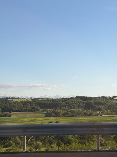 The image depicts a landscape view in Vakarel, Bulgaria. The sky is predominantly clear and blue. To the right, there is a lush green landscape consisting of trees, meadows, and what appears to be cultivated fields. A roadside barrier is present on the right side of the image, giving the impression of looking out from a roadside. In the distance, faintly visible, are possibly snow-capped mountain peaks.