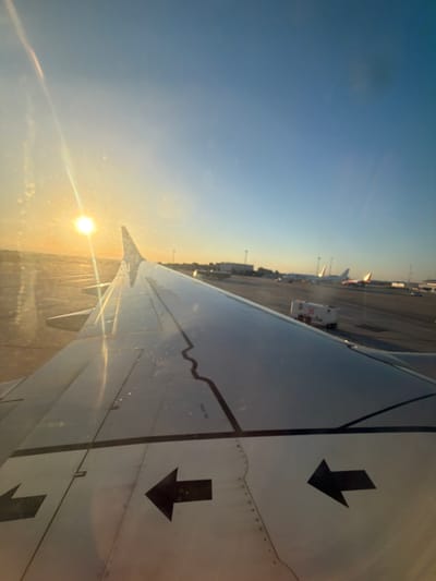 The image is a low-angle, outdoor shot taken from the perspective of an airplane window, looking down at the wing and the tarmac. The time of day appears to be sunrise or sunset, as indicated by the warm, golden light illuminating the scene and the sun's glare. The sky is a gradient of blue, transitioning to a hazy orange near the horizon.
The primary subjects are the airplane's wing, which dominates the right side of the frame, and the airport tarmac visible in the background. The wing is metallic and appears to have some markings, including two black arrows pointing downwards and some faint text that is illegible. The tarmac shows various airport infrastructure, including parked airplanes, service vehicles, and airport buildings. The overall setting is an airport, likely at Cagliari-Elmas Airport (SUF) in Italy, given the provided location context.
The activity taking place is implied to be an aircraft preparing for takeoff or having just landed, as the plane is on the ground. There are no people visible in the frame. The weather appears clear with some atmospheric haze, contributing to the soft lighting. The mood conveyed is one of travel and anticipation, with the warm light suggesting the start or end of a journey.