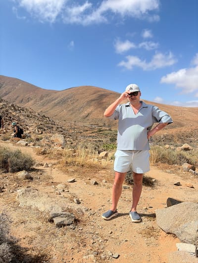 The image depicts an arid, rocky mountainous landscape in Pájara, Fuerteventura, Spain, under a clear blue sky with scattered white clouds.
In the foreground, a Caucasian male, appearing middle-aged, stands on a light brown dirt path. He is wearing a light grey/blue short-sleeved collared shirt with a V-neck, light grey/blue shorts, a light grey baseball cap with a dark triangular logo, dark sunglasses, and dark grey slip-on shoes with lighter soles. His right hand is raised to touch the brim of his cap, while his left hand rests on his hip, holding a multicolored patterned cloth or item tucked into his pocket. He is looking towards the viewer with a slight smile.
To the left in the mid-ground, a second individual, a female, is visible. She is crouching or sitting on the rocky ground, wearing a dark top, dark bottoms, a light blue/green cap, and a black backpack. She is facing away from the primary subject and towards the left.
The ground is predominantly light brown earth, interspersed with numerous rocks and boulders of varying sizes. Sparse, dry, scrub-like vegetation and clumps of straw-colored grass are scattered across the terrain.
The background features multiple undulating, treeless mountains and hills with a reddish-brown hue, showing signs of erosion. A narrow track or road is visible winding along the slope of the furthest hill to the left. In the mid-ground, nestled within a valley between the hills, there is a small area with some greener vegetation, possibly cultivated, and vertical posts suggesting fencing or a structure.
The scene is brightly lit by direct sunlight, casting distinct shadows to the right of objects and individuals, indicating a high sun position.