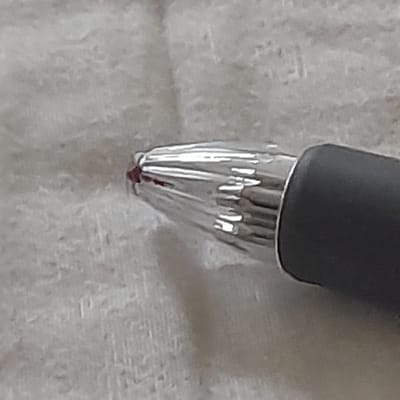 The image shows the tip of a grey and silver pen lying on a white surface. The pen is made of metal and has a red ink tip. The pen is lying on a white surface that appears to be a piece of fabric. There is no other context or information available about the scene or the location.