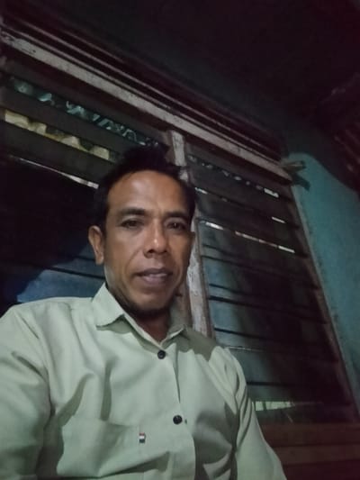 A medium close-up selfie shows a man in a light green collared shirt. He has dark hair and is looking at the camera with his mouth slightly open. The background appears to be a window with wooden blinds and a teal wall. The image is dimly lit, suggesting it was taken indoors at night. The man's shirt has a small pocket on the left side with a red, white, and blue emblem. There are two dark buttons on the shirt. The overall scene is simple and undramatic.