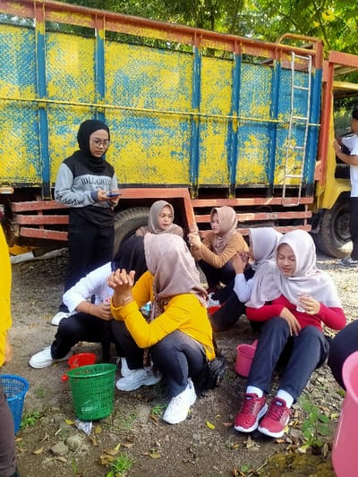 The image captures an outdoor scene in Menjing, Indonesia, featuring a group of individuals gathered near a large, weathered truck. The ground is unpaved, composed of dirt and gravel, with scattered dry leaves and sparse green vegetation. In the background, a large cargo truck is visible, featuring a yellow cab and a cargo bed with heavily chipped yellow paint revealing a blue diamond-plate pattern beneath. A silver metal ladder is attached to the side of the truck's cargo bed.
Seven individuals are present. Six are identifiable as females, and one male is partially visible on the far right. Most of the females wear hijabs. A standing female in the left-center, wearing a black hijab, glasses, a grey and black long-sleeved shirt with "BILLABONG" text, and black pants, holds a smartphone with both hands.
In the foreground and mid-ground, five females are seated or crouching on the ground. One female, wearing a yellow long-sleeved top, black pants, and a light brown patterned hijab, crouches with her hands clasped together near her chest. Adjacent to her, a female in a red long-sleeved top, dark pants, and a light grey hijab sits, holding a small white carton. Another female, obscured behind the yellow-shirted individual, wears a white long-sleeved top and dark pants, also crouching with clasped hands. Two other females are present further back: one in a light brown hijab and top, and another in a dark brown hijab, partially obscured.
Objects visible on the ground include a green plastic basket, a partially visible blue plastic basket on the far left, and a pink plastic bucket on the right. The male individual on the far right is partially visible, wearing a white t-shirt, standing next to the truck. The individuals appear to be engaged in a casual group gathering or break.