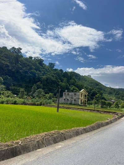 The image captures a scenic rural landscape in Thuận Hòa Commune, Vietnam, characterized by vibrant green rice paddies and a lush, tree-covered hillside under a bright blue sky dotted with fluffy white clouds. A paved road with a concrete drainage ditch runs along the right side of the frame, separating the viewer from the expansive rice field. In the middle ground, a partially constructed or abandoned two-story house with a yellow facade and grey concrete elements stands against the backdrop of the verdant hill. Scattered trees and smaller structures are visible on the lower slopes of the hill. Utility poles with visible wires are present in the mid-ground, suggesting the presence of infrastructure within this rural setting. The overall scene conveys a sense of tranquility and natural beauty common to agricultural regions in Vietnam.