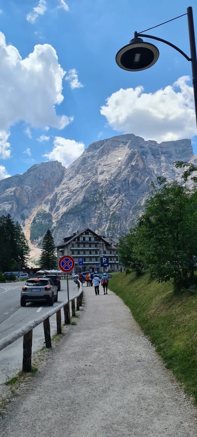 The image depicts a scenic view of the Dolomites in Prags - Braies, Italy. A towering, rugged mountain dominates the background, its rocky face sculpted by time and weather. In the foreground, a paved pathway runs alongside a grassy embankment, bordered by wooden posts.

Several people are walking on the pathway, appearing to be tourists enjoying the landscape. They are casually dressed, suggesting a leisurely outdoor activity. A modern building, possibly a hotel or restaurant, is situated at the base of the mountain, its architecture blending with the natural surroundings.

A street lamp is visible on the left side of the image, its design utilitarian. A car is parked on the side of the road, its brake lights illuminated. Various road signs are present, including a "no parking" sign with a red circle and cross, and parking signs with a blue "P". The sky is a vibrant blue, dotted with fluffy white clouds, indicating pleasant weather. The scene suggests a daytime setting, likely late morning or early afternoon, given the lighting. The overall atmosphere is one of tranquility and natural beauty.