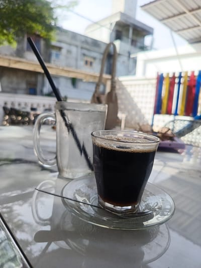 The image depicts an outdoor table scene in the city of Pantegajah, Indonesia. In the foreground, two glass beverages are positioned on a light-colored, reflective table surface. A small, clear glass tumbler on the right contains a dark liquid, presumably coffee, topped with a thin layer of light brown foam or crema. This tumbler rests on a clear glass saucer. To its left, a larger, clear glass mug is filled with ice cubes and water, containing a black plastic straw. A thin, black, rod-like object, likely a stirring stick, lies across the coffee saucer. The table surface reflects the light and surrounding objects, showing subtle variations in tone and texture.

The midground is out of focus but reveals an urban environment. On the left, a portion of a grey building wall with an obscured window and some green foliage is visible. On the right, a structure with vertical colored bars (red, yellow, blue) stands prominently, possibly part of a fence or decorative element. Additional blurry objects, including a light-colored bag, are situated in the background, suggesting other patrons or personal belongings.

The background is heavily blurred, displaying various indistinct architectural structures and a bright, clear sky, consistent with an outdoor setting under daylight. No individuals are present in the frame.