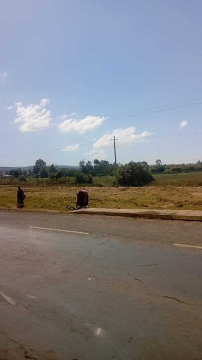 The image depicts an outdoor scene under a clear blue sky with scattered white clouds. The setting appears to be a rural or semi-rural area alongside a paved road.
In the background, a landscape of rolling hills or elevated ground is visible on the left, covered with dense green vegetation, possibly trees or forests. Towards the center and right, the background transitions to flatter, open fields with patches of green grass and some dry, yellowish-brown vegetation. Scattered bushes and trees are interspersed across this distant landscape.
The mid-ground features a wide expanse of dry, light-brown grass and dirt, with some greener patches. A tall, slender wooden utility pole stands upright near the center-right of the image, with power lines or communication cables extending horizontally across the sky.
In the foreground, a paved asphalt road occupies the lower portion of the frame. The road surface is grey, showing signs of wear with cracks and some faint white road markings visible on its left and right edges. A light-colored concrete curb separates the road from the grassy verge.
Two individuals, appearing to be women, are present on the grassy area next to the curb.
One individual, on the left, is standing facing away from the camera, looking towards the right. They are dressed in a long, dark-colored garment, possibly a wrap or dress, with some lighter patterns or colors visible on the back. They appear to be carrying something or have an item draped over their left arm.
The second individual, on the right, is crouching or kneeling close to the ground, also with their back mostly turned towards the camera. They are wearing a dark red or maroon garment, possibly with some detailing. This person seems to be engaged in an activity on the ground, possibly tending to items or sorting something, as some indistinguishable objects are visible near their hands on the grass.
The lighting suggests bright daylight, likely mid-morning to late afternoon, given the distinct shadows and overall brightness. No text is visible in the image.