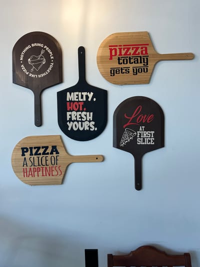 This image shows five wooden pizza peels mounted on a white wall. From left to right, top to bottom, the pizza peels display the following text and graphics:

*   **Top Left:** A dark brown pizza peel with white text that reads "NOTHING BRING PEOPLE TOGETHER LIKE PIZZA" encircling a graphic of a heart pierced by two arrows and wrapped in a ribbon.
*   **Top Center Right:** A light-colored wood pizza peel with red and black text. The text reads "pizza totally gets you". The word "pizza" has a red underline, and the words "totally" and "gets you" have black underlines.
*   **Center Left:** A black pizza peel with white and red text. The text reads "MELTY. HOT. FRESH. YOURS." "HOT." is in red.
*   **Center Right:** A dark brown pizza peel with red and white text and a graphic of a slice of pizza. The text reads "Love AT FIRST SLICE". The word "Love" is in red script, and the other text is in white. The slice of pizza is white.
*   **Bottom Left:** A light-colored wood pizza peel with black and red text. The text reads "PIZZA A SLICE OF HAPPINESS". "PIZZA" and "A SLICE OF" are in black, and "HAPPINESS" is in red.

The wall appears to be a plain white surface. Part of a dark wood chair back is visible at the bottom of the image.

The location of this image is not provided in the prompt.