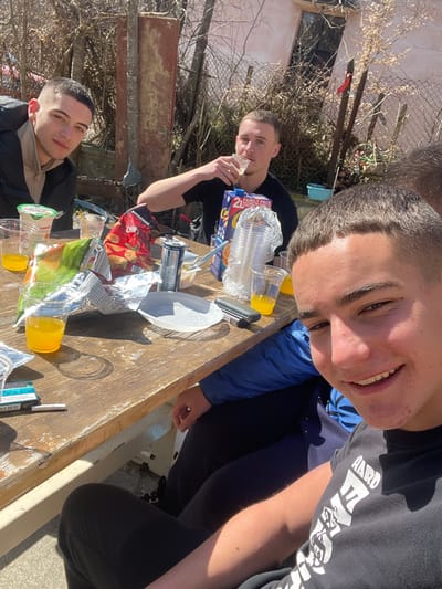 The image captures four young males gathered around a weathered wooden picnic table outdoors in Kisyovtsi, Bulgaria, under direct sunlight.
In the foreground, a male with short dark hair and a black t-shirt featuring white text smiles directly at the camera. To his immediate left, another male's legs are visible under the table, clad in black pants and a blue jacket. On the opposite side of the table, to the left, a third male with short dark hair, wearing a black jacket over a light-colored top, looks towards the camera with a neutral expression. In the center-background, a fourth male, also with short dark hair and a black t-shirt, holds a clear plastic cup to his mouth, actively drinking.
The wooden table is laden with remnants of a casual gathering. Visible items include multiple clear plastic cups, some containing orange-colored liquid, others empty; an open bag of "Knuspride" brand potato chips; an open carton of "Cappy" brand orange juice; a stack of unused clear plastic cups; several aluminum beverage cans (one with a prominent blue top); a white plastic plate; and a turquoise-blue cigarette pack, likely Parliament Aqua Blue, with a single cigarette and a black lighter resting beside it.
The background reveals a sparse outdoor setting. Bare tree branches and a wire fence are visible behind the table, suggesting a late winter or early spring season. A light-colored building with a dark window is partially seen further back. The ground beneath the table is a mix of dirt and scattered debris. Strong shadows are cast by the individuals and objects, indicating bright sunshine.