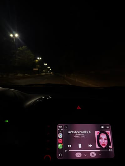 The image shows the interior of a car at night, with the dashboard visible. The car is presumably in motion, as suggested by the blurred streetlights seen through the window on the left. A digital display screen is mounted on the dashboard, showing a music player interface with the song "LUCES DE COLORES" by Omar Courtz and Primera Musa playing. The screen also displays icons for various apps and the time, 11:35 PM. The emergency light button is illuminated. The environment is dark, with only the dashboard and the streetlights outside providing illumination. The location of Tuxtla Gutiérrez, Mexico, is not directly identifiable from the image but can be inferred based on the language settings of the music player being in Spanish.