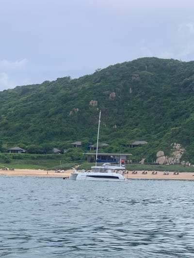 The image captures a panoramic view of a coastline in Vinh Hai, Vietnam, under an overcast sky. In the foreground, calm blue water reflects the muted light. A white catamaran sailboat is prominently positioned in the mid-ground, its mast reaching towards the sky. Behind the boat, a sandy beach stretches along the shore, lined with lounge chairs and a few scattered umbrellas, suggesting a resort or recreational area. Several low-lying buildings with dark roofs are nestled amongst the trees on the gentle slope leading up to a densely forested, green hillside. Rocky outcrops are visible on the hillside, and a few people can be seen on the beach. A small, narrow boat with a yellow sail is also present near the shore. The overall scene conveys a sense of tranquility and a tropical, coastal environment.