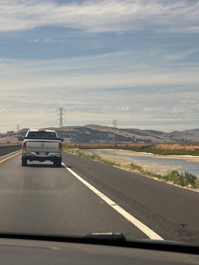A white Dodge Ram pickup truck is driving on a highway. The truck is in the left lane of a two-lane highway, and the camera is positioned behind the truck, giving a rear view. The truck has a license plate that appears to be from California. The highway is asphalt and has a white solid line marking the left edge of the lane. To the right of the highway is a body of water, possibly a marsh or estuary, with vegetation along the banks. In the distance, rolling hills are visible with power lines and transmission towers scattered across the landscape. The sky is partly cloudy with a mix of blue sky and white clouds. The scene appears to be in a rural or semi-rural area.