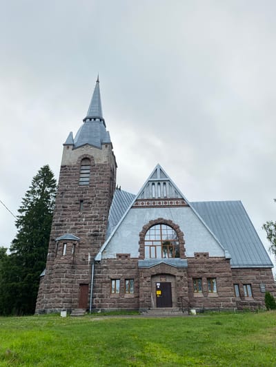 The image shows a stone building with a tall, pointed steeple, likely a church, located in the city of Melnikovo, Russia. The building's facade is constructed with large, rough-hewn reddish-brown stones, interspersed with lighter-colored stone accents. A gray, sloped roof covers the main structure, while the steeple is also capped with a gray, conical roof. Several windows are visible on the building, some with decorative stained glass. A large arched window with a grid pattern is prominently featured above the main entrance. The area in front of the building is a grassy slope with scattered small white flowers. The sky above is overcast and gray. A tall evergreen tree stands to the left of the building.