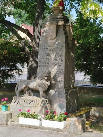 This image captures a stone war memorial located in Jesenice, Czechia, likely in a park or public garden, on a sunny day. The central subject is a tall, light-colored stone monument, partially shaded by large, leafy trees. At the base of the monument stands a stone statue of a roaring lion, resting on a carved rock formation.

The monument's main visible panel is engraved with numerous names, including "CHANDEL VAC," "GLICK JOS," "PROUAZNIK BED," "LOUZESKY KAR," "STYCH KAREL," "NEULIDA FR.," "RATAJ KAREL," "CHUDOMEL ST," and "DUORAK LAD." Below these names, a dedication reads: "SUYM DRAHYM VOJINUM PADLYM VE SVETOVE VALCE 1914-1920 VENUJI OBČANÉ JESENSTI." This translates to "To our dear soldiers who fell in the World War 1914-1920. Dedicated by the citizens of Jesenice." Another side panel also lists names like "RECHKA JOS," "SAZIMA VAC," and "BLAZEK FINT."

Flower pots containing pink and red geraniums are placed at the base of the memorial, adding a touch of vibrancy to the solemn structure. The bright sunlight casts clear shadows from the surrounding trees and the monument itself. In the background, partially obscured by dense foliage, a building with a reddish-brown roof can be seen, along with a metal fence. The scene conveys a sense of remembrance and peace.