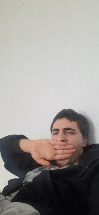 A fair-skinned person with dark hair is positioned in the upper right corner of the image, turned sideways. They are covering their mouth with their hand. They are wearing a dark jacket over a light-colored shirt. The background consists of a stark white wall occupying the majority of the left side and extending behind the individual. The image appears grainy and low-resolution.