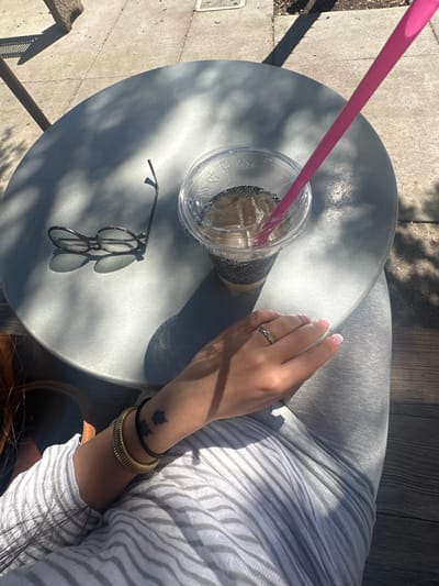 An overhead view captures an outdoor scene at a circular, grey metallic table in Glendale, United States. On the table's right side, a clear plastic cup containing an iced beverage, presumably coffee, with a black patterned sleeve and a clear lid, has a pink plastic straw inserted. To the left of the cup, a pair of dark-framed eyeglasses rests with one arm extended. Dappled sunlight casts distinct shadows across the table surface. A person's right arm and hand are visible in the lower right portion of the frame. The hand displays a silver-toned ring with a patterned band on the ring finger and a French tip manicure. The wrist is adorned with a gold-toned ribbed cuff bracelet and a thin black band. A small, dark-inked tattoo, appearing as two stylized figures, is present on the inner forearm. The person wears a light-colored, horizontally striped top and light grey fitted pants. A brown shoe is partially visible on the far left. The ground visible beyond the table is concrete pavement.