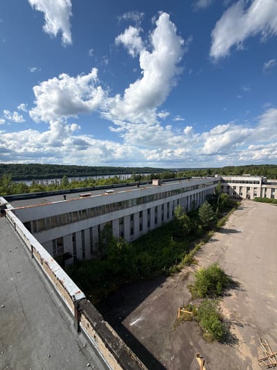 The image shows a long, multi-story abandoned industrial building, likely in or near Nikolsky, Russia. The building has a flat roof and a facade made of white brick with rows of windows. Many windows are broken or boarded up, and vegetation is growing around the building and on the rooftop. The surrounding area appears to be an overgrown industrial site with a paved courtyard showing signs of disrepair and debris. In the background, a body of water, possibly a river or lake, is visible, bordered by a dense forest. The sky is blue with scattered white clouds. There are no people or active elements visible in the scene.
