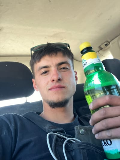 A young man is holding a bottle of "ICKO" lemonade in a car. He's wearing a dark-colored hoodie and has sunglasses perched on his head. The interior of the car is light-colored. The location is unknown.