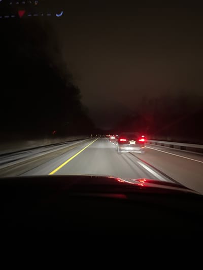 A nighttime scene shows a highway viewed from a vehicle. The road is lined with guardrails and illuminated by red taillights fading into the distance. A yellow dividing line is visible. To the left, a dark and foggy landscape merges into the blackness of night. The right side of the image is mostly dark, likely the interior of the vehicle.