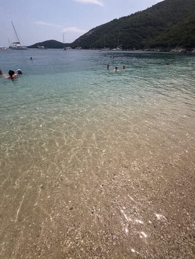 The image captures a clear, shallow bay in the Municipal Unit of Ellomenos, Greece, characterized by its pristine turquoise waters lapping onto a pebble beach. The seabed, visible through the translucent water, is a mosaic of small stones in various shades of brown and grey, illuminated by dappled sunlight.

In the foreground, the water is so clear that the texture and color of the pebbles beneath the surface are distinct. As the water deepens towards the middle ground, it transitions to a richer blue. Several individuals are seen enjoying the water. In the left of the frame, a group of people are partially submerged, with some appearing to be swimming or wading. Further out, scattered individuals are visible swimming and floating in the bay.

In the background, several sailboats are anchored in the calm waters, their white hulls and tall masts contrasting with the deep blue of the sea and the lush green of the hillside that slopes down to the water's edge. The hillside is densely covered with dark green vegetation, suggesting a Mediterranean climate. The sky above is a pale blue with scattered, wispy white clouds. The overall impression is one of a serene and picturesque coastal setting on a sunny day.