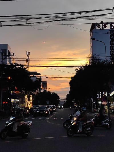 The image captures a busy city street at dusk in Hòa Cường Ward, Vietnam. A sunset featuring an orange-yellow horizon transitions into a grey upper sky, providing the primary light source from the background. Numerous black electrical and communication wires are strung horizontally across the top section of the frame.

The foreground depicts an asphalt road with visible dashed white lane markings. Multiple motorcycles, primarily scooters, are on the road, with headlights illuminated. Individuals operating these vehicles wear helmets. One white scooter is prominent on the left in the foreground, with its rider wearing light trousers. Further down the road, several other motorcycles and a few dark-colored cars are visible, indicating active traffic.

On the left side of the street, multi-story buildings are silhouetted against the sky, featuring illuminated storefront signs with Vietnamese script. A red traffic light is visible on this side, and a cellular tower structure is present atop one of the buildings. Trees with dense, dark foliage line the street on both sides.

On the right, a multi-story building displays vertical signage reading "SISA" and "BA HUNG." A street light stands adjacent to this building. Additional illuminated storefronts are visible further down the right side of the street, interspersed with tree canopies. Vehicles appear to be moving or queuing along the roadway.