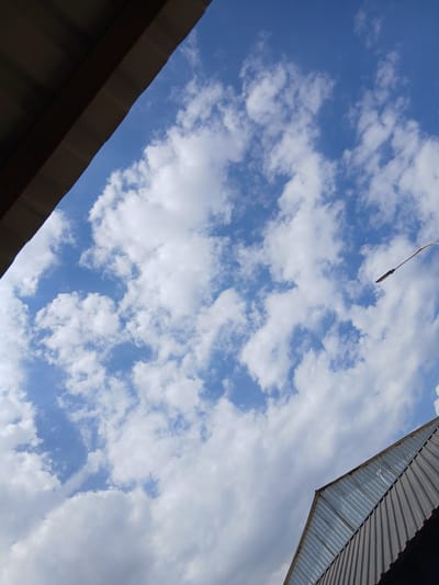 The image shows a view looking up at a blue sky with scattered white clouds. Two parts of a building's roof are visible in the foreground, one in the upper left corner and another in the lower right. The roof in the upper left appears to be dark and corrugated. The roof in the lower right is also corrugated and appears to have sections of glass or translucent material. A portion of a street lamp or utility pole is visible in the upper right quadrant of the image, extending diagonally upwards. The scene is outdoors, and the sky suggests daytime. The location is identified as being in the city of Kanchoch, Tajikistan.